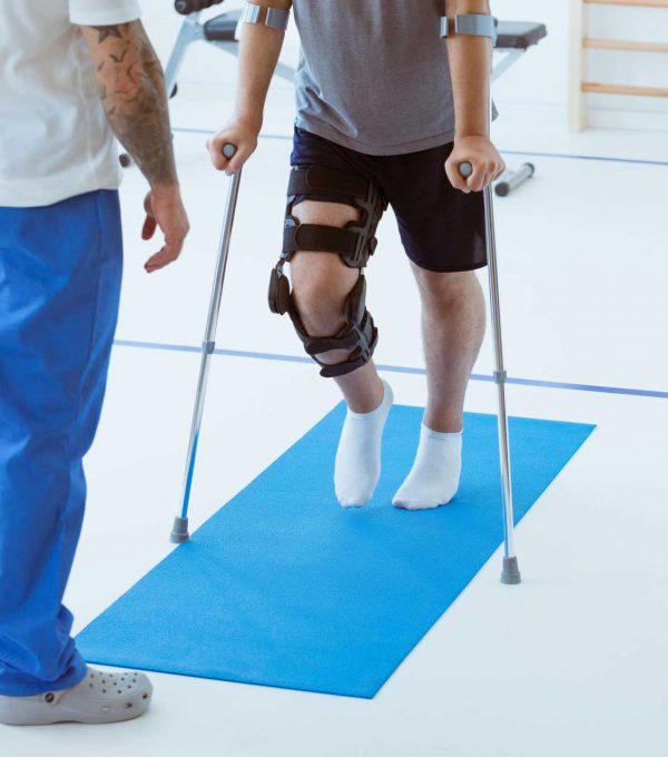 Man after car accident in an orthosis and on crutches learning to walk in the clinic, helpful therapist near him